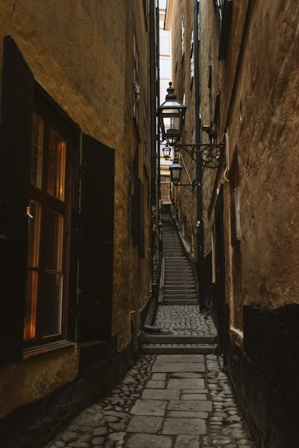 A narrow cobbled alleyway between two weathered, yellowish-brown buildings with dark-framed windows, leading uphill towards a fire escape staircase. The buildings' exterior walls show signs of aging and wear, with some areas painted black at the base. Metal brackets and small lamps are attached to the walls, and the stone pathway is uneven with larger paving stones near the edges. The staircase at the top is made of metal, with steps and railings visible, indicating a typical urban residential setting. This outdoor scene captures the tight space often encountered during house relocations in historic or densely built areas, where careful fitting and transportation of furniture or boxes might be required. The lighting suggests daytime with natural light diffusing down the alley, aligning with moving and packing processes handled by companies like Man with Van Lambeth, especially in locations such as Camberwell.