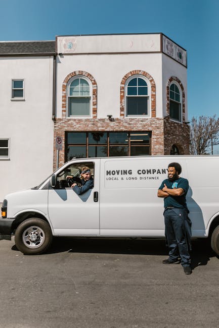 A man with a beard and wearing a black cap, white t-shirt, and jeans is loading three large cardboard boxes into the side of a white commercial van parked on a residential street. The van, equipped fo