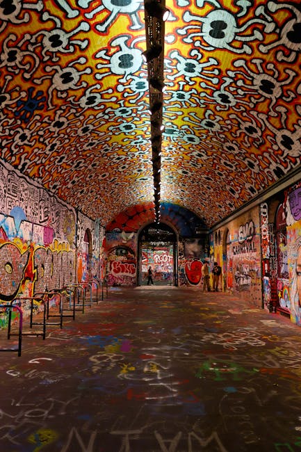 A long, arched underground tunnel decorated with colorful graffiti art on all walls and the ceiling, featuring abstract patterns, faces, and various tags. The tunnel is illuminated by natural light at the far end, with a black pipe running along the center of the ceiling. The floor is covered with spray paint markings, and several metal benches are positioned along the left wall. The scene depicts an urban setting potentially used for street art or photography, with the entrance at the far end visible. This setting is inside a tunnel that may be part of a cityscape or transit network, and the vibrant artwork provides a visually striking backdrop relevant to urban environments associated with house removals or relocation services.