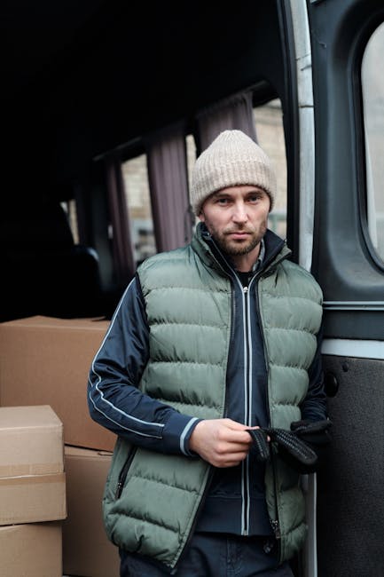 A man with a serious expression is standing next to the open side door of a moving van during a home relocation. He is wearing a beige knitted beanie, a grey puffer vest over a dark blue jacket with white piping, and black gloves. Inside the van, visible are several large brown cardboard boxes stacked neatly, likely containing household items or packing materials. The interior of the vehicle is illuminated by natural light filtering through the windows. The background shows a residential street with pavement, suggesting the loading or unloading process is taking place outside a home. The scene reflects a typical furniture transport and packing process involved in professional house removals, with the man prepared for the loading process as part of a relocation service offered by Man with Van Lambeth.