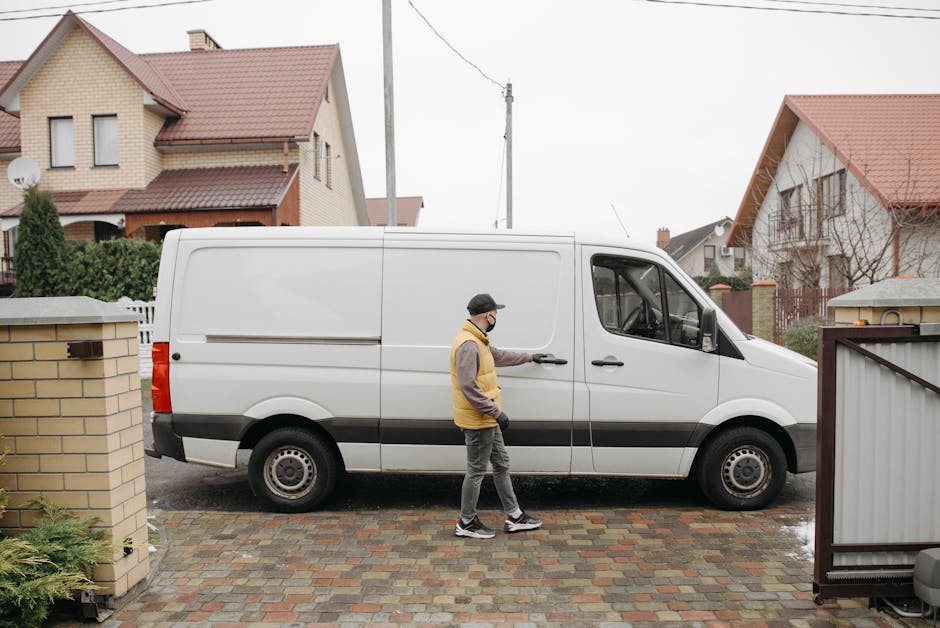 A man wearing a face mask, beige and grey jacket, and grey joggers is seen standing next to a white cargo van parked on a paved driveway in a residential area. The van, part of a home relocation or furniture transport process, is positioned parallel to a brick wall and a closed metal gate, with the driver’s door open. In the background, there are several detached houses with pitched roofs, some with terracotta tiles, and a utility pole with overhead wires. The scene appears to be outdoors during daytime with overcast weather, and the man is either preparing for or in the midst of loading or unloading items from the van, which is equipped for moving services by Man with Van Lambeth. The environment includes modest garden hedges and a paved street surface, supporting a typical house removal setting involving packing materials and furniture ready for transport.