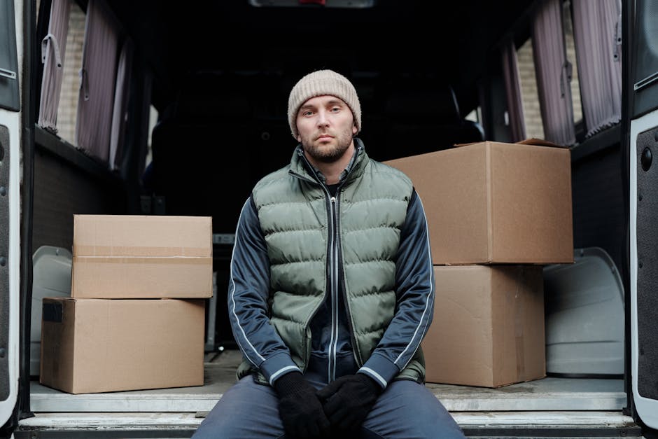 A man with a serious expression is standing next to the open side door of a moving van during a home relocation. He is wearing a beige knitted beanie, a grey puffer vest over a dark blue jacket with white piping, and black gloves. Inside the van, visible are several large brown cardboard boxes stacked neatly, likely containing household items or packing materials. The interior of the vehicle is illuminated by natural light filtering through the windows. The background shows a residential street with pavement, suggesting the loading or unloading process is taking place outside a home. The scene reflects a typical furniture transport and packing process involved in professional house removals, with the man prepared for the loading process as part of a relocation service offered by Man with Van Lambeth.