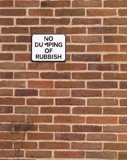 A rectangular white sign with black text attached to a red brick wall, displaying the message 'NO DUMPING OF RUBBISH' in bold capital letters. The sign is positioned towards the upper left side of the
