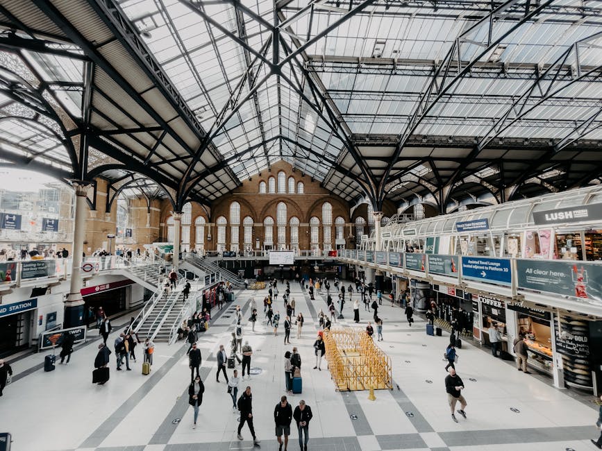 Interior view of a busy train station with a high arched glass roof supported by black metal framework, allowing natural light to illuminate the spacious concourse. Numerous travelers are walking across the tiled floor, some carrying luggage, while others stand or sit by the commercial stalls and seating areas along the edges. On the right, a row of retail shops and kiosks, including a LUSH store, are visible with signage and displays. In the center, a group of people are moving towards escalators leading to an upper level, where additional staircases and seating areas are present. A yellow trolley with empty stacks of luggage or boxes is positioned near the bottom right, suggesting a scene involving packing, unpacking, or home relocation activities. The scene captures the dynamic environment typical of a major station, with the atmosphere indicative of transit and logistics in a moving or relocation context, as supported by services from Man with Van Lambeth.