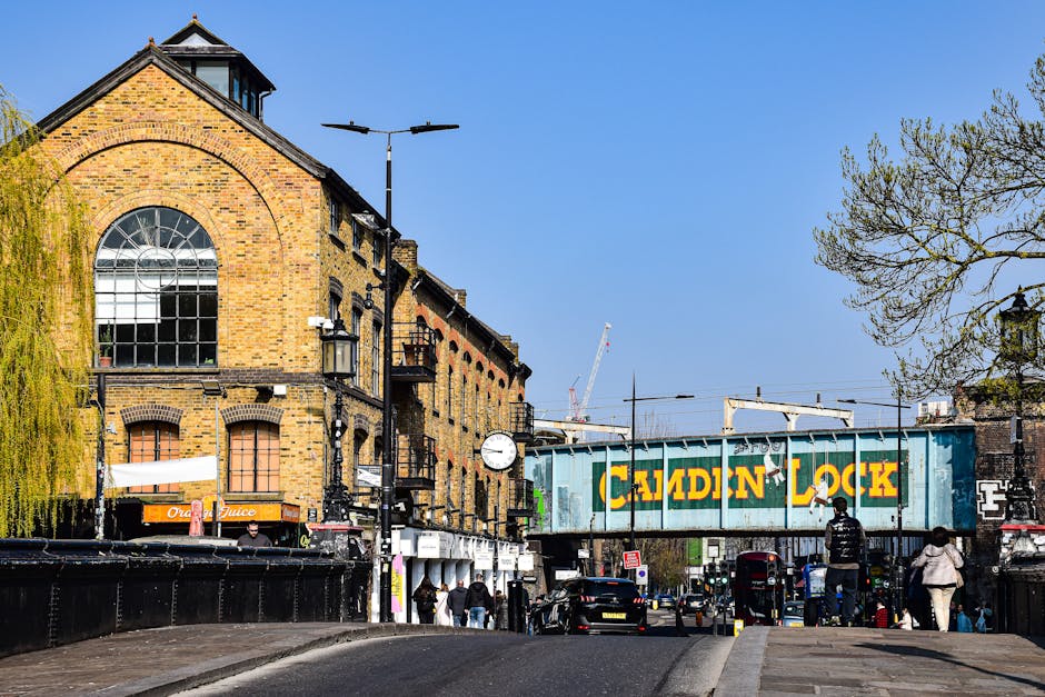 A street scene in Lambeth featuring a historic brick building with large arched windows and a clock on the exterior wall, situated alongside a railway bridge painted with a green and yellow 'Caledonian Lock' sign. In the foreground, pedestrians walk along the pavement on a clear day with blue skies, and cars are parked or moving on the road. The surrounding environment includes leafless trees and street lamps, with construction cranes visible in the background. The scene depicts urban transportation and movement, consistent with home relocation activities and furniture transport in the Lambeth area, reflecting the typical setting for professional removals services such as those provided by Man with Van Lambeth.