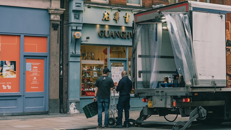 Two men dressed in casual clothing are positioned on the pavement outside a storefront during a home relocation process, with one man holding a small hand truck and the other carrying a plastic crate. They are preparing to load furniture or boxes into a large, open-sided moving truck parked on the street. The truck has a partially transparent plastic curtain on its side, revealing a stack of cardboard boxes and packing materials inside. The storefront behind them displays Asian-style signage with gold lettering and the name 'GUANGH' visible on the window, which also shows various items and promotional materials. The scene is captured in natural daylight, highlighting the urban setting typical of house or apartment moves involving furniture transport and packing during a loading process. The logistics of home removals carried out by firms like Man with Van Lambeth are implied through the context of loading and packing materials visible in the image.