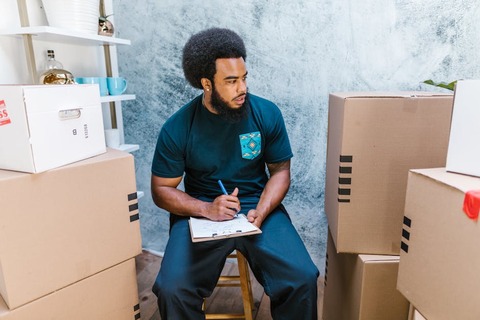 A young man with dark skin and curly black hair, dressed in a navy blue t-shirt with a patterned pocket, is seated on a small wooden stool inside a room with a textured gray wall. He is holding a blue pen and taking notes on a clipboard resting on his lap while surrounded by numerous cardboard moving boxes of various sizes, some sealed with packing tape and others open. The boxes are stacked on the floor and on a white shelving unit in the background, which also holds decorative items like a small plant, glasses, and a ceramic container. The room is well-lit, suggesting a home or apartment space preparing for a house move, with visible activity related to packing and organizing, related to house removals and furniture transport services provided by Man with Van Lambeth.
