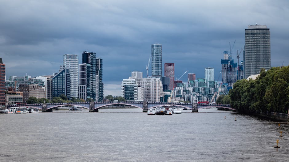 A wide view of the South Bank area in London with the City of London skyline visible in the background, featuring numerous modern high-rise buildings and construction cranes. In the foreground, the River Thames is shown with several white boats and a few moored vessels, and a bridge crossing the river connects the two sides. On the right, a line of lush green trees runs alongside the riverbank, providing a natural contrast to the urban environment. The scene is illuminated by natural daylight under a partly cloudy sky. This image captures the bustling city landscape typical of London’s skyline, relevant to house removals and furniture transport services provided by Man with Van Lambeth as part of their home relocation and moving logistics operations.