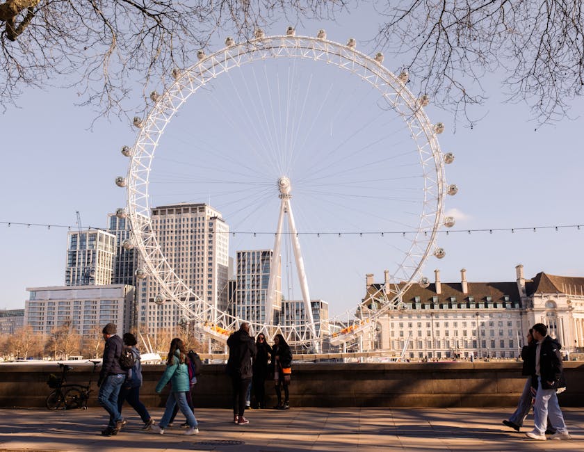 A wide view of the South Bank area in London with the City of London skyline visible in the background, featuring numerous modern high-rise buildings and construction cranes. In the foreground, the River Thames is shown with several white boats and a few moored vessels, and a bridge crossing the river connects the two sides. On the right, a line of lush green trees runs alongside the riverbank, providing a natural contrast to the urban environment. The scene is illuminated by natural daylight under a partly cloudy sky. This image captures the bustling city landscape typical of London’s skyline, relevant to house removals and furniture transport services provided by Man with Van Lambeth as part of their home relocation and moving logistics operations.