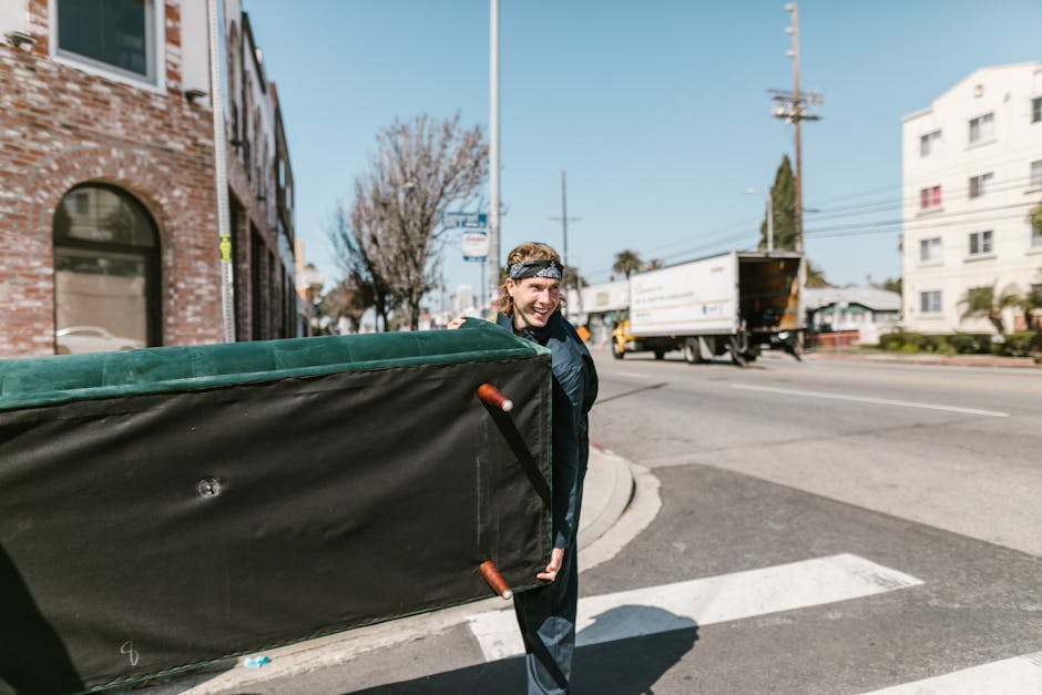 A man with a headband and a dark jacket is outside on a city street during daylight, carrying a large, rectangular piece of furniture wrapped in black moving blankets with orange handles, which he is holding at one side. In the background, a white delivery truck with its rear door open is parked on the road, and several multi-story residential buildings, including a red-brick building with arched windows and a white building, are visible along the street. There are also trees, utility poles, and power lines evident in the scene, indicating an urban environment suitable for home relocation or furniture transport. The visible pavement and street markings suggest a well-maintained area, supporting the logistics of packing and moving tasks in house removals, as associated with professional moving services provided by Man with Van Lambeth.