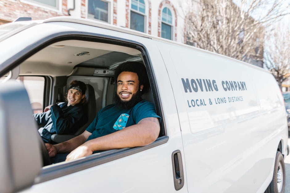 Inside a white moving van with the sliding door open, two professional removalists are visible, seated in the front cabin. The driver, a man with dark, curly hair and a beard, is smiling at the camera, wearing a blue [COMPANY_NAME] branded uniform. The passenger, a woman with dark hair tied back, is also smiling and dressed in a similar uniform, indicating team coordination in a home relocation or furniture transport process. The interior of the van shows visible equipment such as straps, blankets, and packing materials prepared for loading. Outside the van, a blurred urban street scene with nearby residential or commercial buildings and leafless trees suggests a city or suburban environment typical of Lambeth. This scene exemplifies a professional house removals operation involving careful packing, loading, and transportation of household items, supporting the comprehensive moving services offered by [COMPANY_NAME].